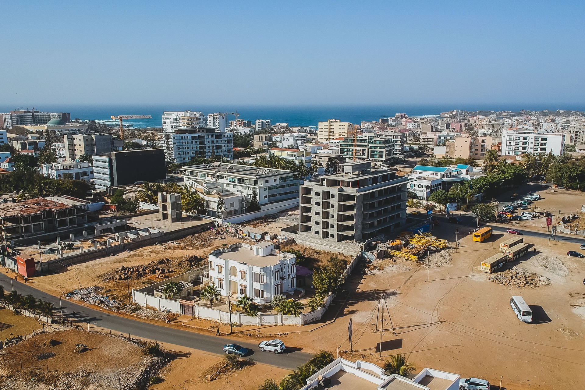 An aerial view of Ngor, Dakar, shows a construction site with cranes and buses near a white villa. The cityscape extends to the ocean, with modern buildings and a clear blue sky, indicating urban growth by the coast.