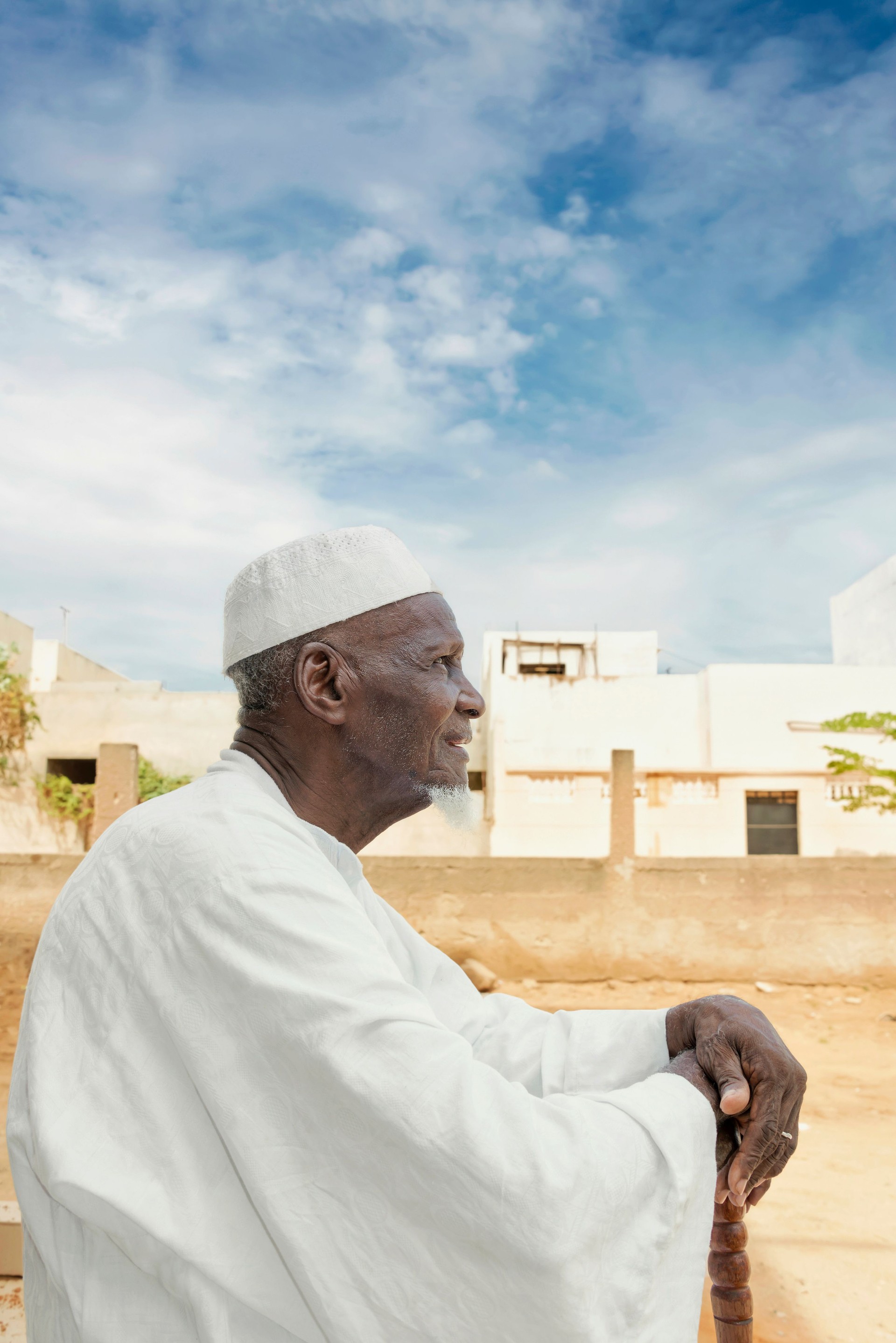 African old man sitting in front of his house and holding a cane, white celebration clothing, eighty years old, photo